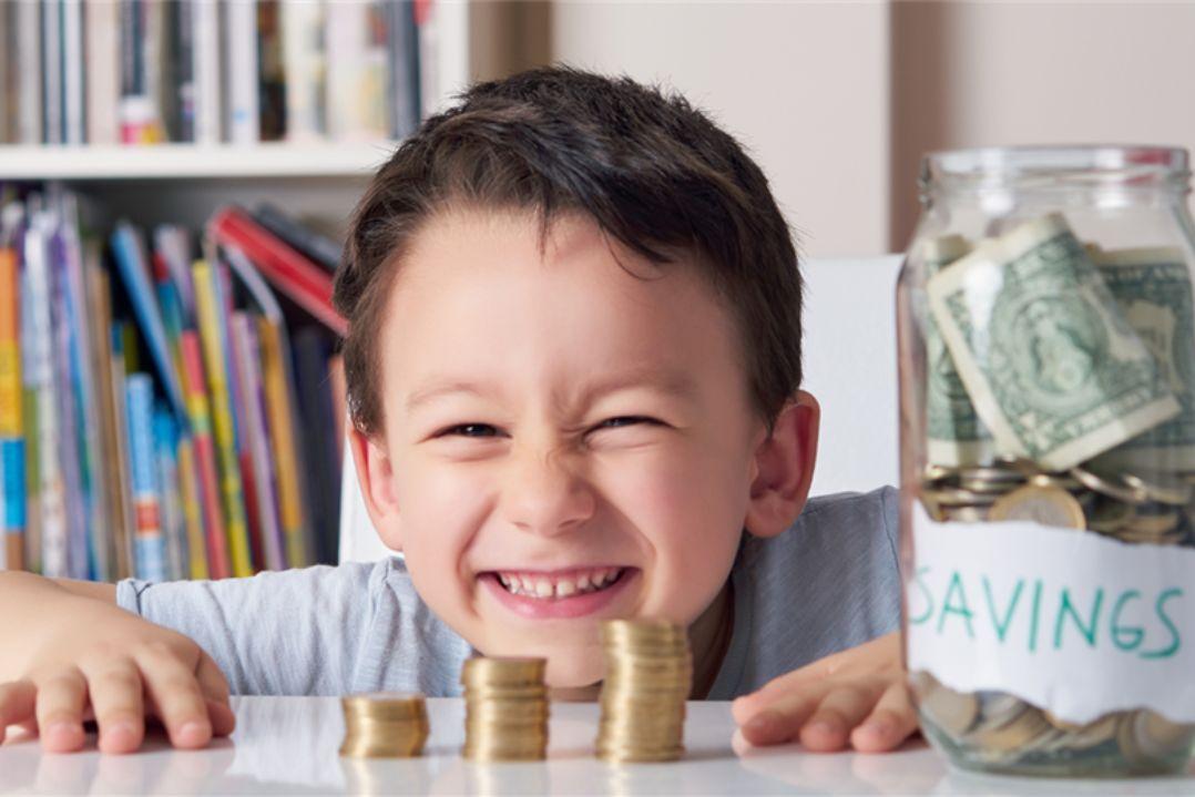 A smiling child counts coins stacked on a table with a savings jar full of cash beside him, conveying joy and financial responsibility. Books are in the background.