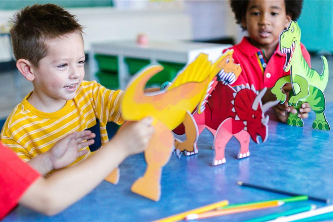 Children play happily at a table with colorful dinosaur toys. The scene is lively and vibrant, conveying joy and creativity in a classroom setting.