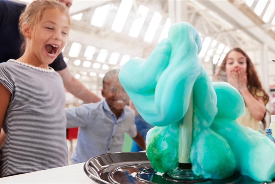 A group of children and an adult watch in awe as a bright blue foam erupts from a science experiment, creating an exciting and energetic atmosphere.