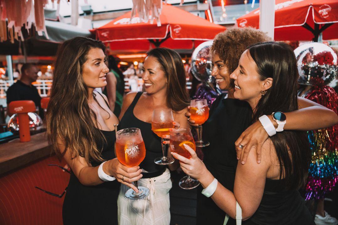 4 woman smiling and hugging while whole a bright orange drink in a glass