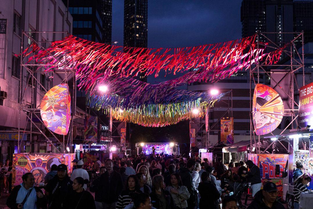 A vibrant street festival at dusk with a colorful canopy of red and purple lights overhead. Crowds gather below, surrounded by high-rise buildings.