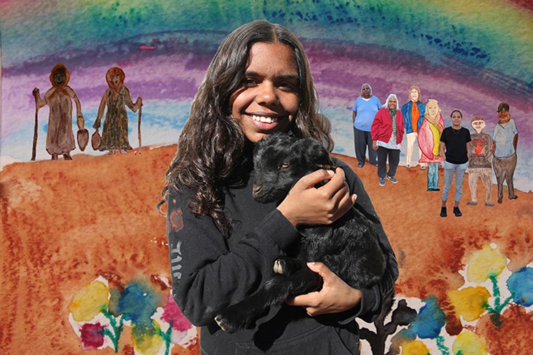 A woman with long curly hair smiles, holding a black dog. Behind her, a colorful mural features a rainbow and people, creating a joyful atmosphere.