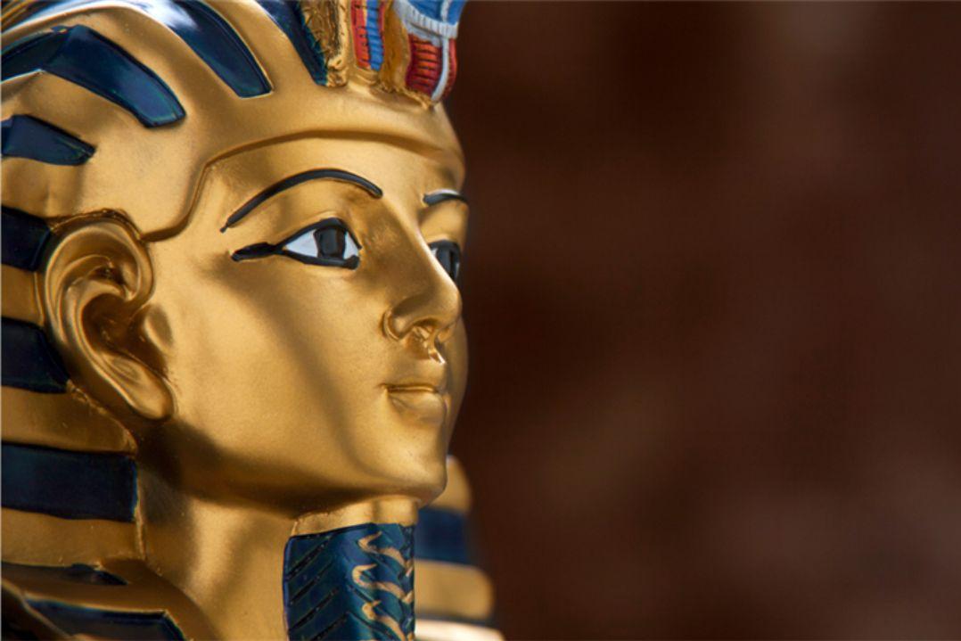 Close-up of an Egyptian pharaoh's golden bust with blue stripes, gazing forward with a regal expression against a blurred brown background.