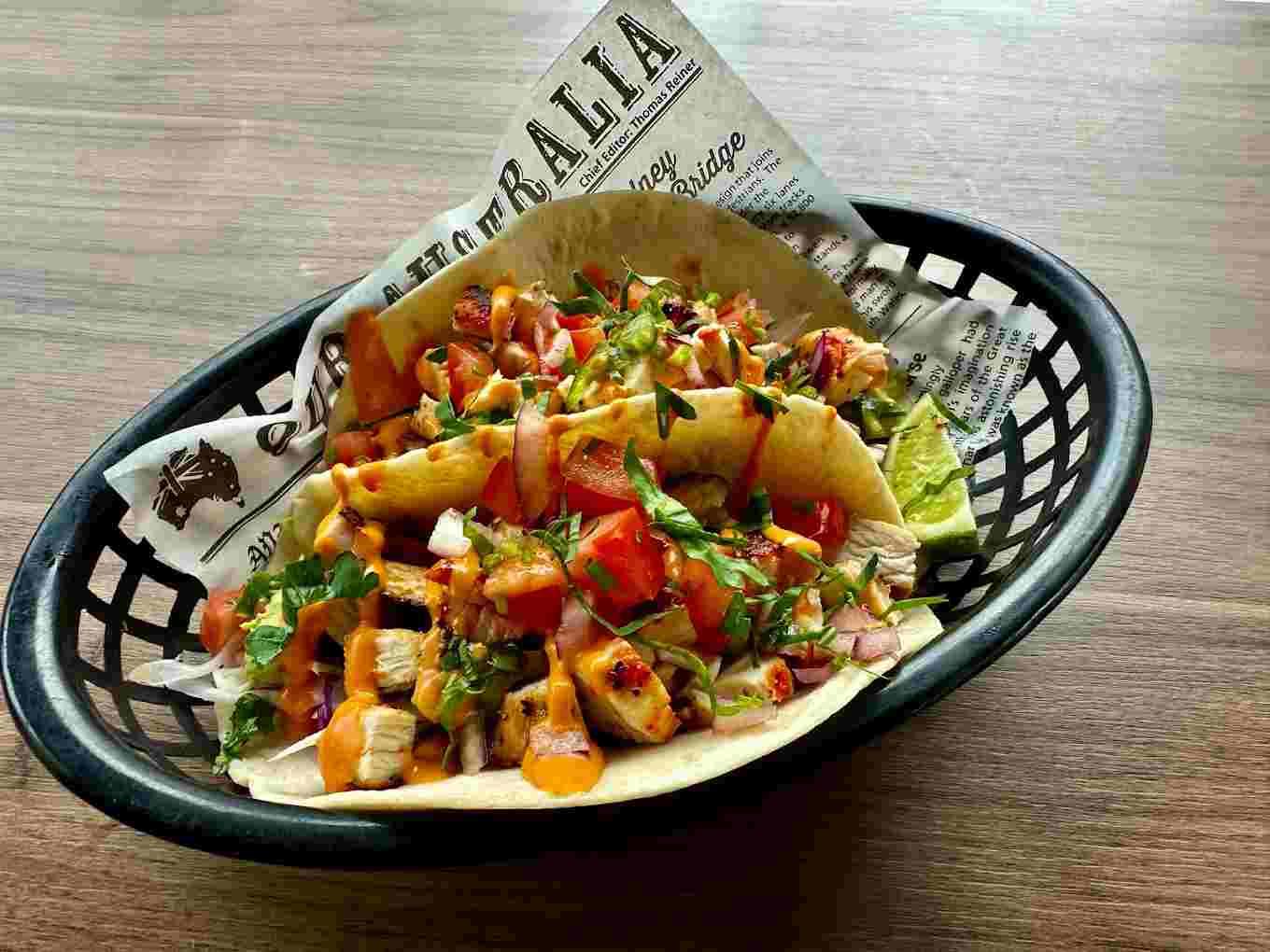 A vibrant taco filled with grilled chicken, diced tomatoes, onions, and fresh cilantro, drizzled with sauce, sits in a black basket lined with newspaper.
