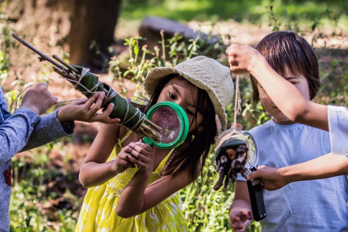 Four children play outdoors, examining plants with magnifying glasses. A girl in a hat peers closely, conveying curiosity and teamwork.