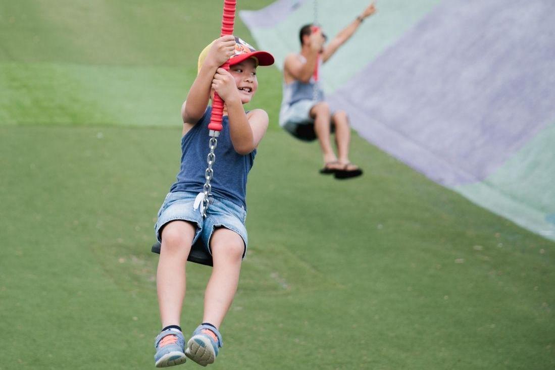 A joyful child in a red cap rides a zip line with a big smile, wearing a denim outfit. A background of a sloped grassy hill sets a playful tone.