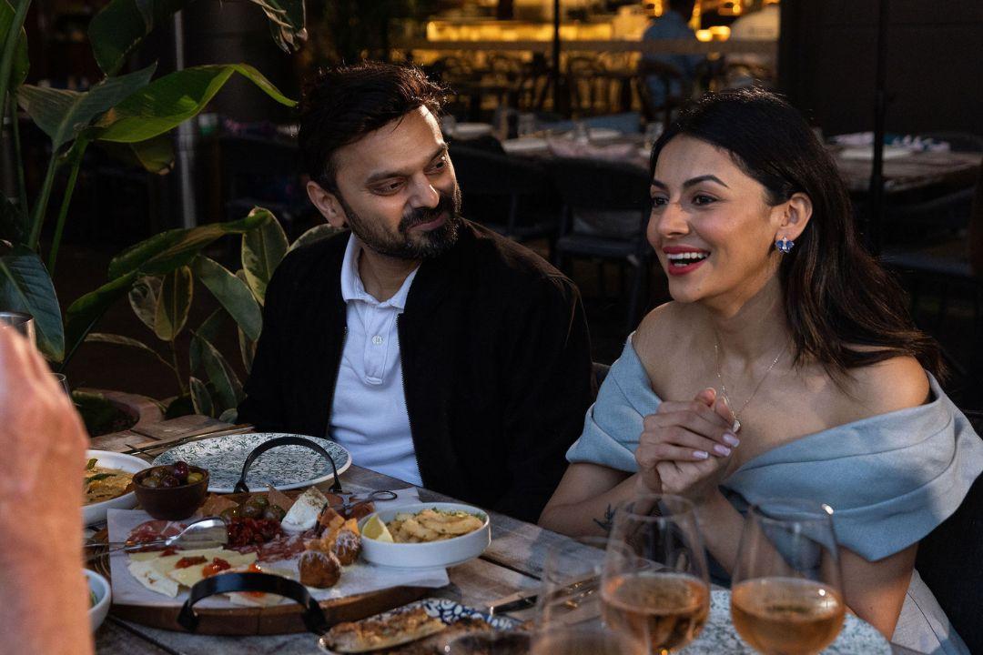 A couple sits at an outdoor table, smiling and engaged in conversation. Warm lighting, greenery, and shared dishes create a cozy, intimate atmosphere.