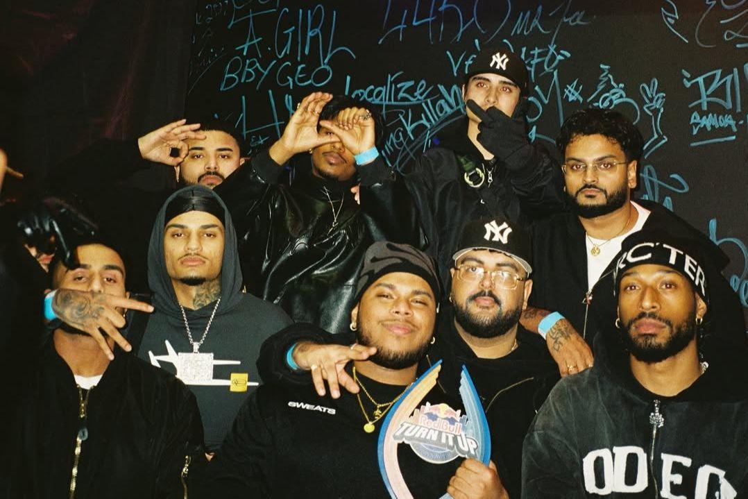 A group of nine people stand closely together, dressed in casual dark clothing. Some make hand signs, while all have neutral to confident expressions.