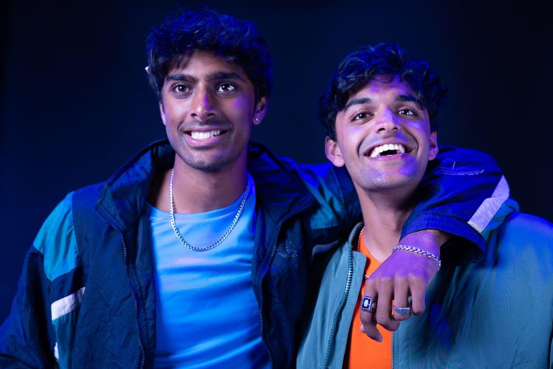 Two young men smiling, one with his arm around the other's shoulder, wearing casual jackets. The lighting is blue, creating a joyful, friendly mood.
