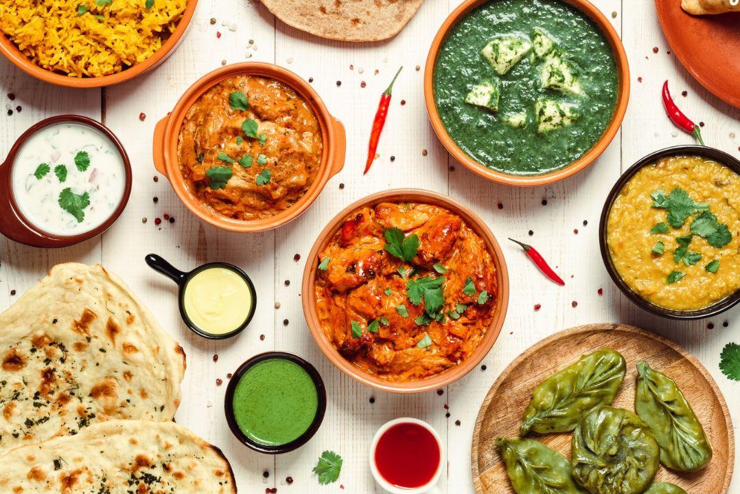 Assorted Indian dishes on a table, including naan, colourful curries, rice, and chutneys. The vibrant setting conveys a rich and flavourful dining experience.