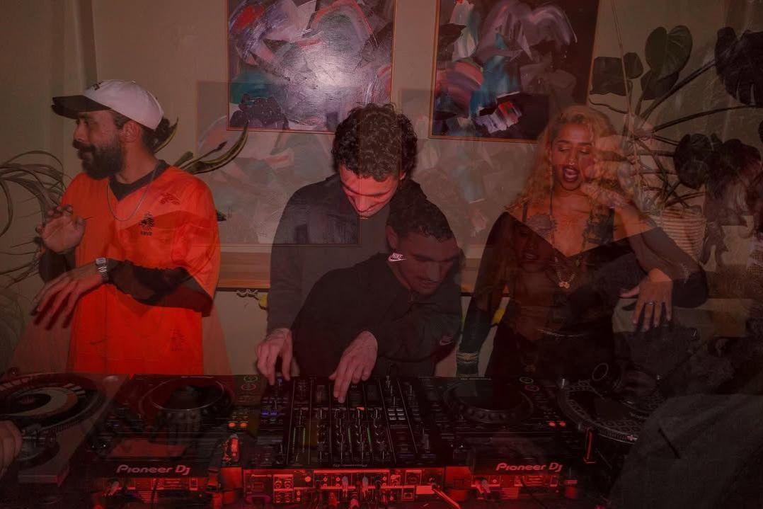 Three DJs are performing in a dimly lit room. The scene is energetic, with colourful lights reflecting off the equipment and posters on the wall.