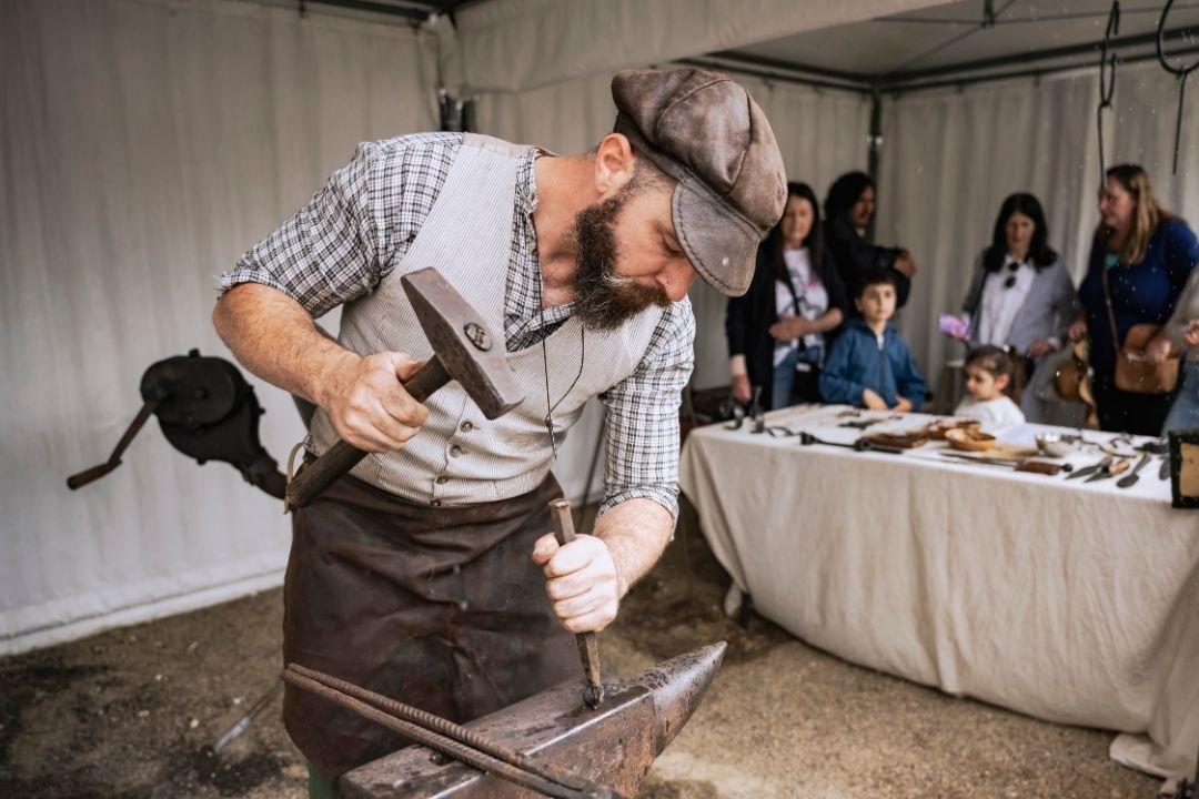A blacksmith in a hat and apron intensely works on metal with a hammer in a studio. Onlookers in the background observe the craft with interest.