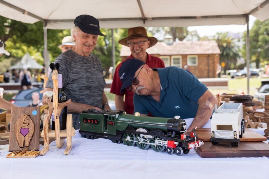 Three people examining a miniature train model on a table at an outdoor craft fair. They appear focused and engaged. Sunlit trees are in the background.
