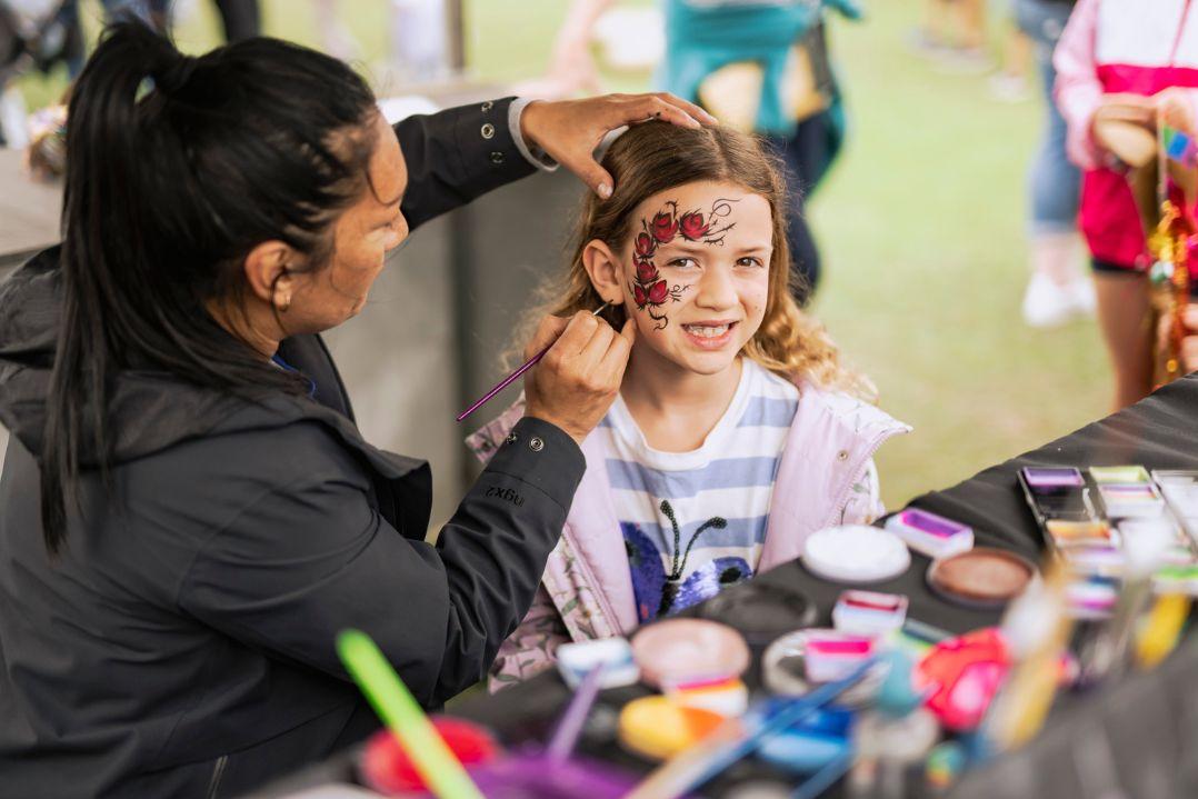 A woman face paints a butterfly on a smiling girl's cheek at an outdoor event. The table is cluttered with colourful face paints, creating a joyful scene.