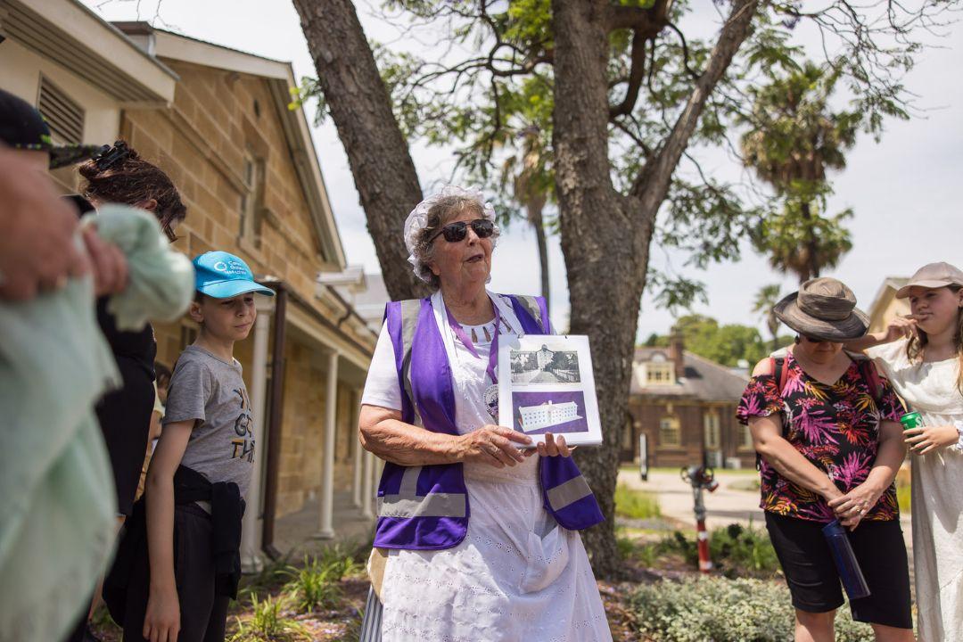 A woman in a white dress and straw hat holds a historical photo, smiling as she speaks to a group outdoors. The mood is educational and engaging.