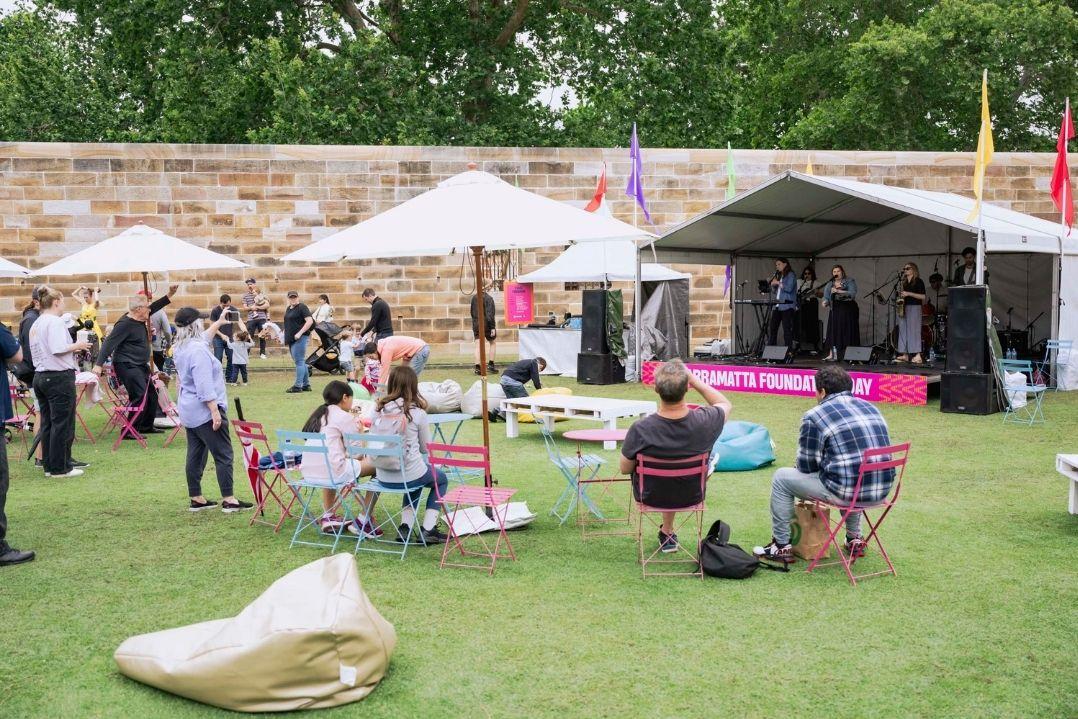 Outdoor music event in a park, people seated on the grass with umbrellas. A band plays on stage under a tent. Relaxed summer atmosphere.