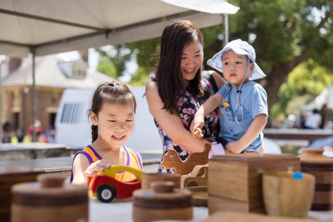 A mother with two young children enjoys a sunny day at an outdoor market. They are smiling while exploring jars of spices on a table, conveying warmth and curiosity.