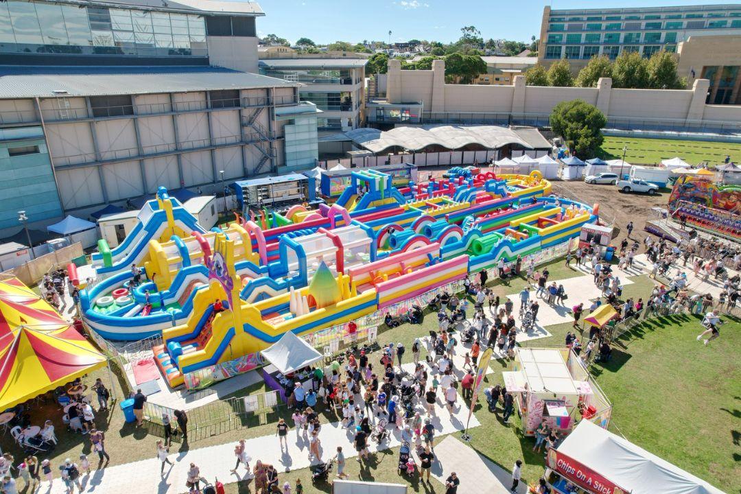 A large, colorful inflatable obstacle course with slides and tunnels, surrounded by a crowd of people enjoying a sunny event. Energetic and vibrant atmosphere.