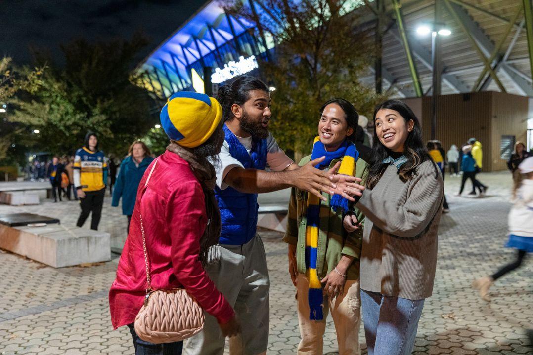 A group of four people stand outside a stadium at night, smiling and chatting. They appear excited, with one holding a sports jersey.