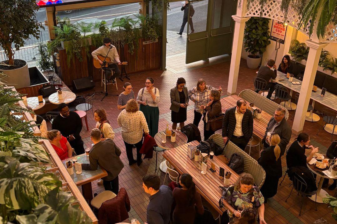 A lively restaurant scene with people mingling around wooden tables. The atmosphere is warm and social, with greenery and soft lighting setting a cozy tone.