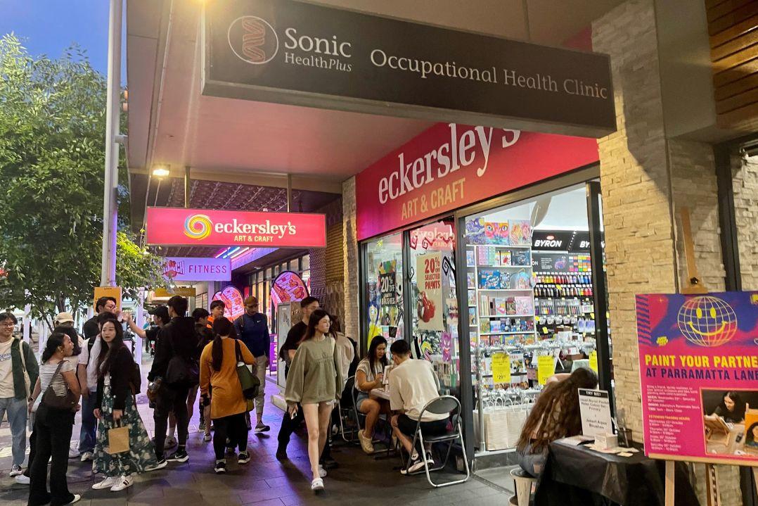A busy street scene at dusk shows people walking past Eckersley’s Art & Craft store with bright signage. A "Paint Your Partner" event is advertised outside.