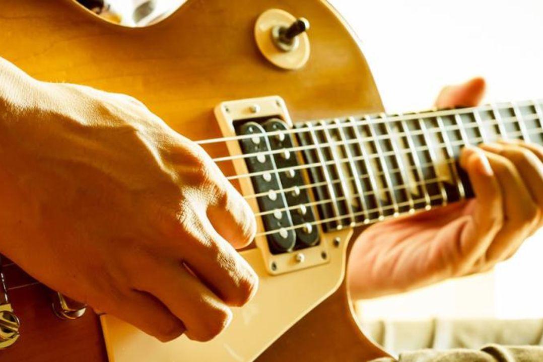 Close-up of hands playing a sunburst electric guitar, focused on the fretboard and strings. The warm lighting creates an energetic and passionate mood.