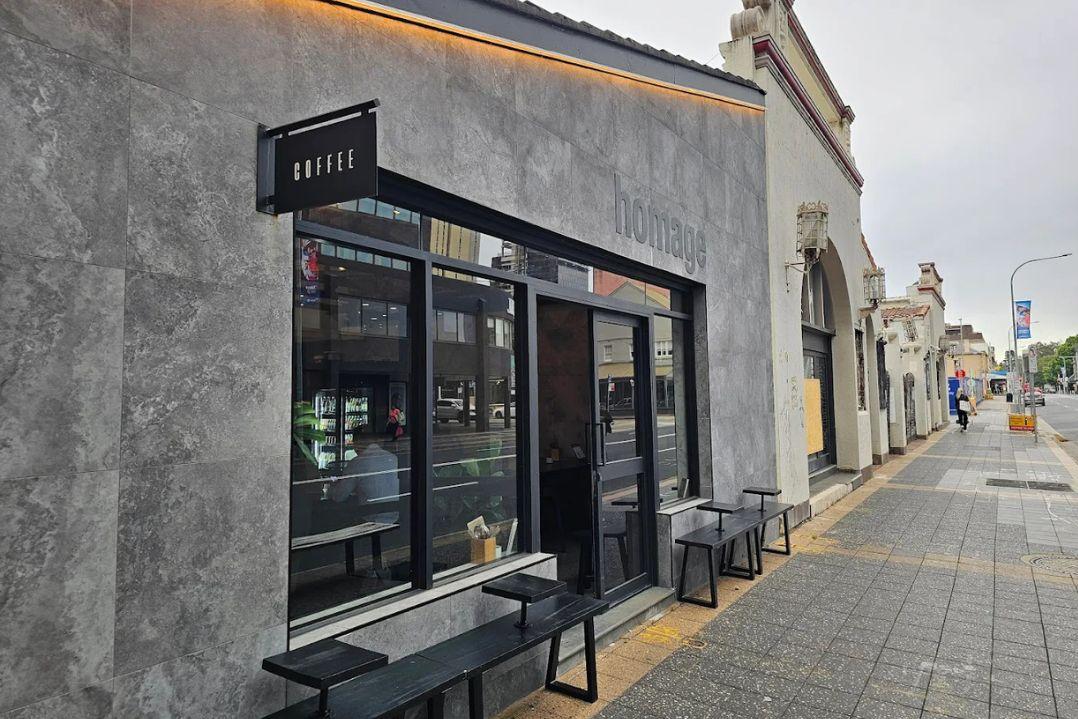 Street view of a modern gray cafe with large windows, black door, and outdoor seating. Overcast sky sets a calm, urban ambiance.