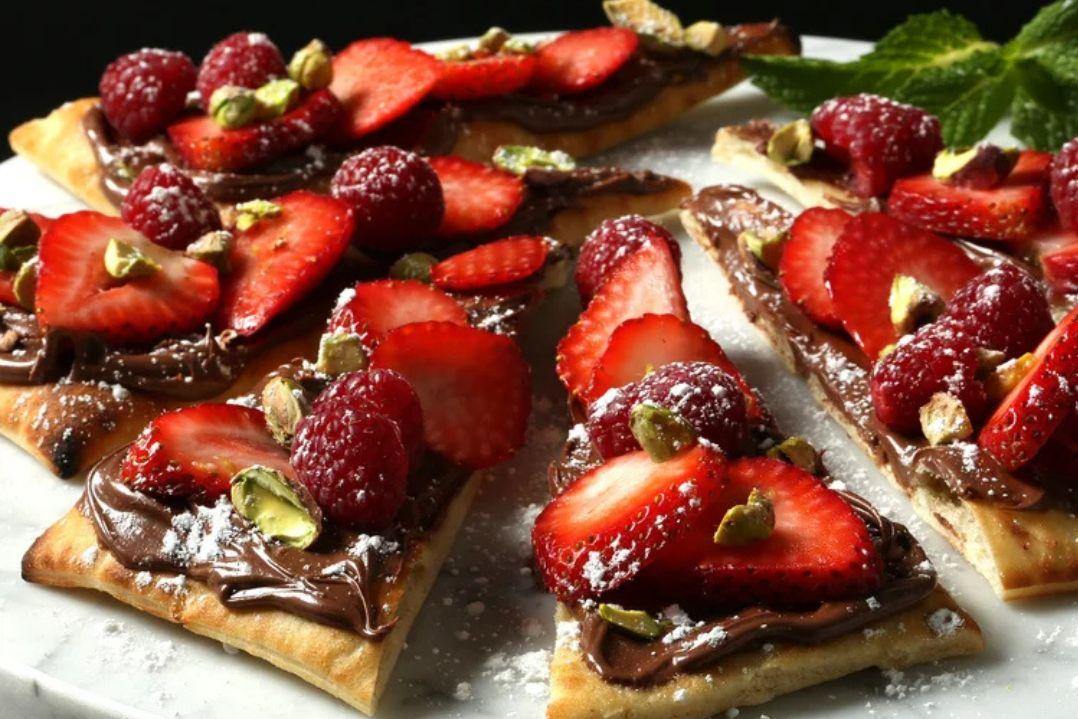 Close-up of dessert pizza topped with Nutella, fresh strawberries, and raspberries on a light surface. The image conveys a decadent and appetizing tone.