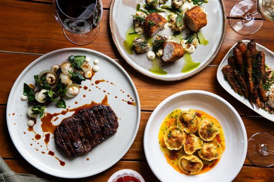 A wooden table with dishes of gourmet food: steak with greens, tortellini in sauce, and a fresh salad. Wine glasses and a cozy ambiance suggest fine dining.
