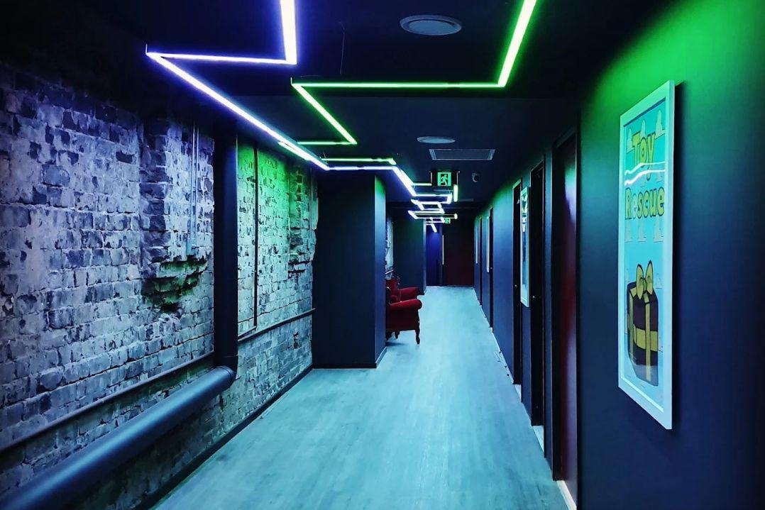 A modern hallway with vibrant green and blue neon lights on the ceiling, textured stone walls, and framed artwork. The atmosphere is sleek and futuristic.