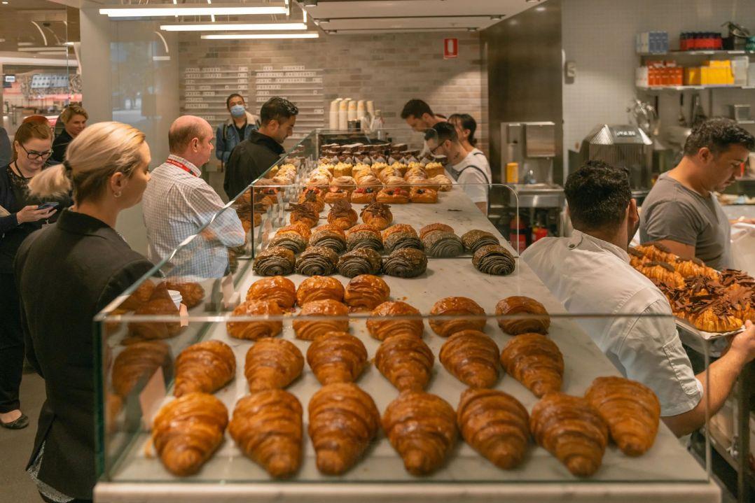 Bakery scene with loaves of fresh bread on racks. Staff in aprons work efficiently amid a warm, bustling atmosphere, evoking freshness and community.