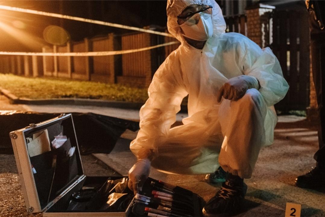 A forensic investigator in a white suit crouches at a nighttime crime scene. They examine tools from an open case near a numbered evidence marker, with police tape in the background.