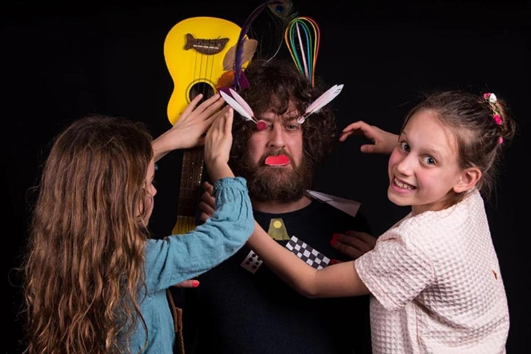 A man with a playful expression stands surrounded by two girls adding quirky decorations like a toy guitar, feathers, and paper cutouts to him. The scene is lively and humorous.