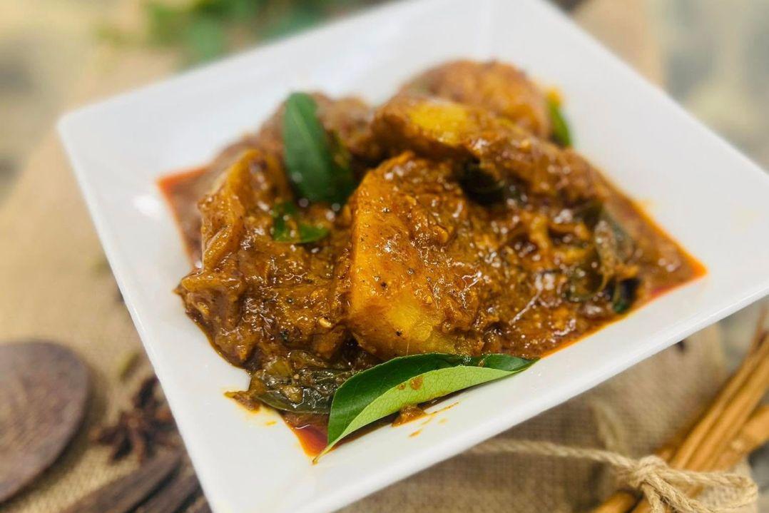 A square white plate holds a vibrant fish curry, featuring pieces of fish in a rich, spicy red sauce garnished with herbs. The setting is warm and inviting.