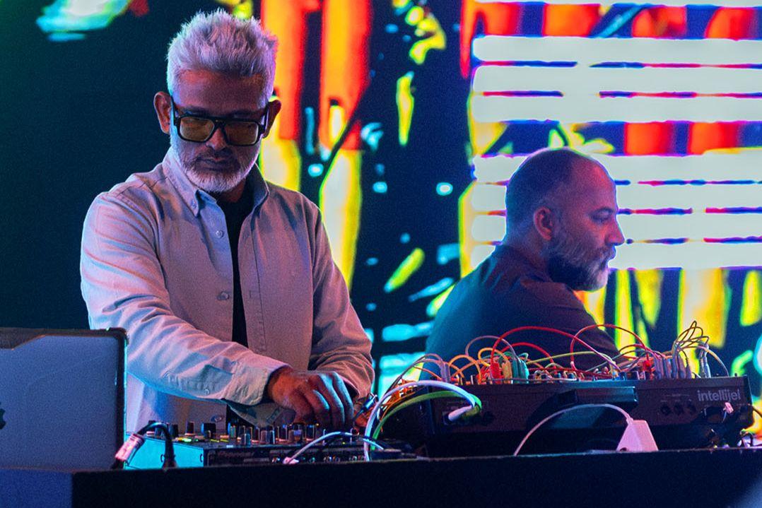 Two DJs perform on stage with vibrant neon lights in the background. One wears a white shirt and tie, the other a dark shirt, both focused and immersed.