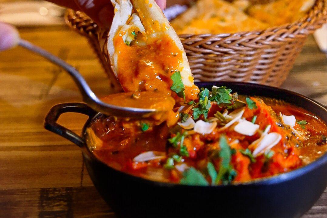 A skillet of vibrant chili topped with cilantro and diced onions. A hand dips a tortilla chip into the chili. A basket of chips is blurred in the background.