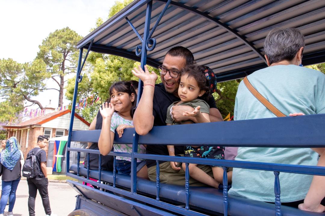 A happy family scene on a blue trolley. A man embraces two children as they wave warmly. Background features trees and a house. Joyful, lively atmosphere.