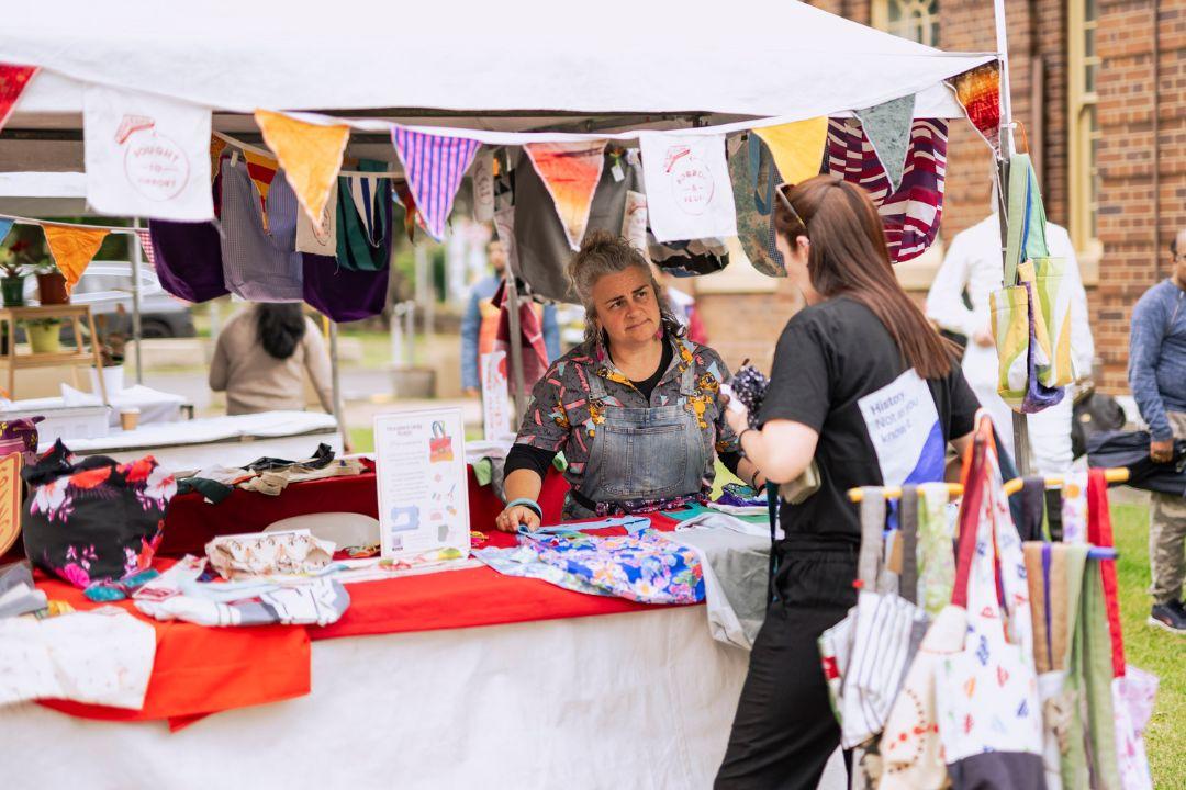 A woman smiles while working at a colourful outdoor market stall with hanging bunting. A customer in a hat looks at various handmade items. Festive atmosphere.