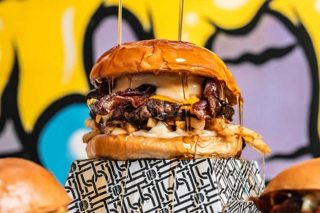 A juicy hamburger with melted cheese and caramelized onions sits atop a patterned surface, backed by a colorful, abstract mural. Bold and appetizing.