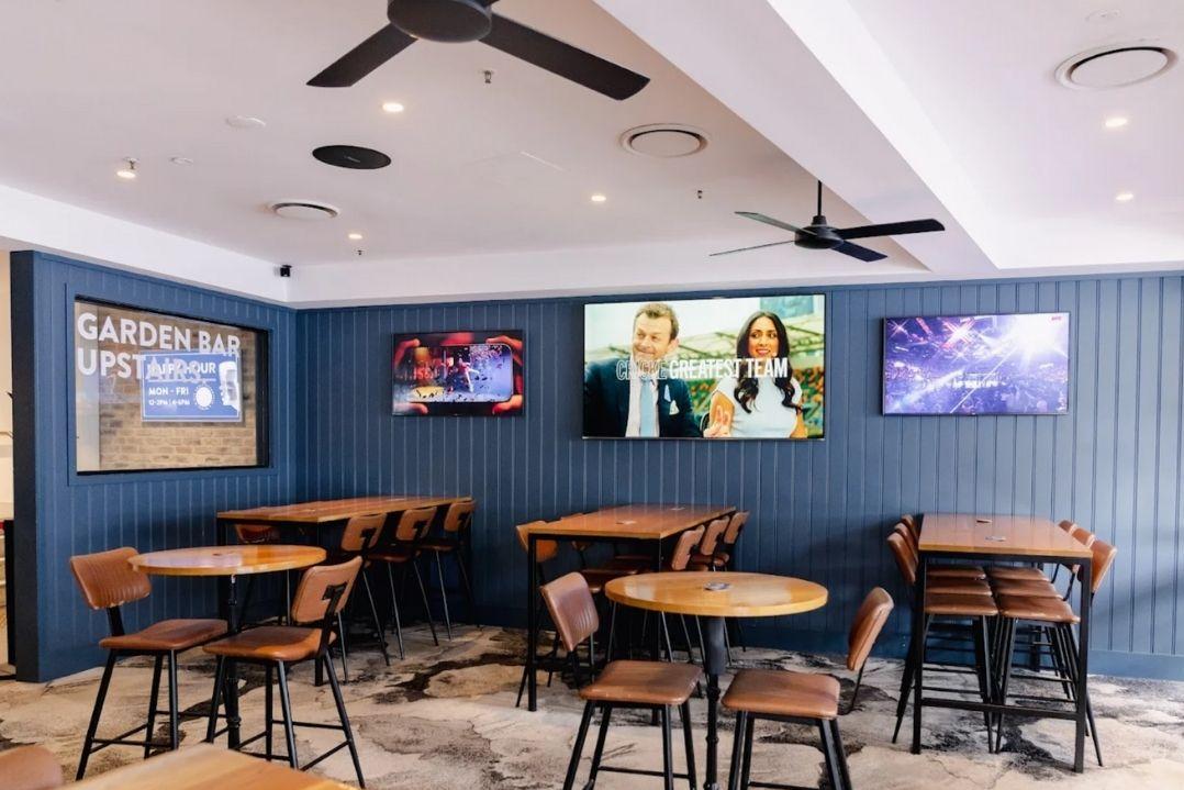 Cozy hotel with wooden tables and chairs under ceiling fans. Three vibrant paintings hang on a navy-blue wall, adding color and a welcoming vibe.