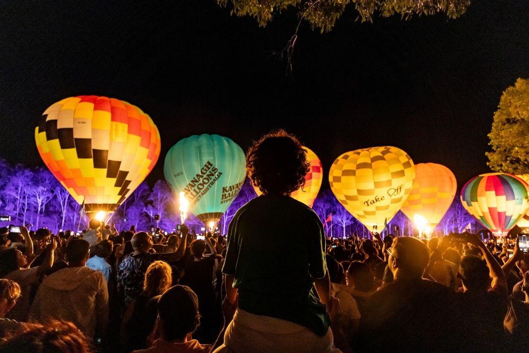A child on a parent's shoulders watches brightly lit, colourful hot air balloons against a night sky, surrounded by a joyful, captivated crowd.