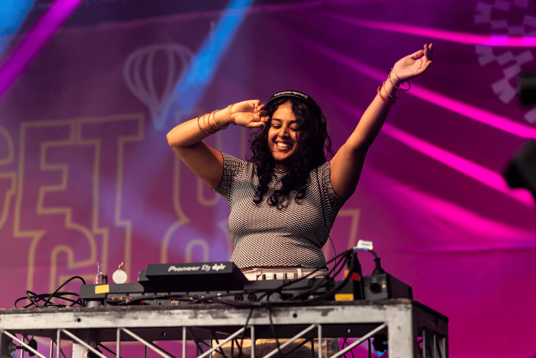 A DJ in a grey shirt energetically performs on stage, with one hand on headphones and the other raised. Vibrant pink and purple lights create a lively atmosphere.