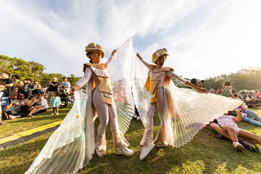 Two performers in shimmering costumes with large, flowing wings dance on grass under a bright sky. Onlookers sit nearby, enjoying the festive atmosphere.