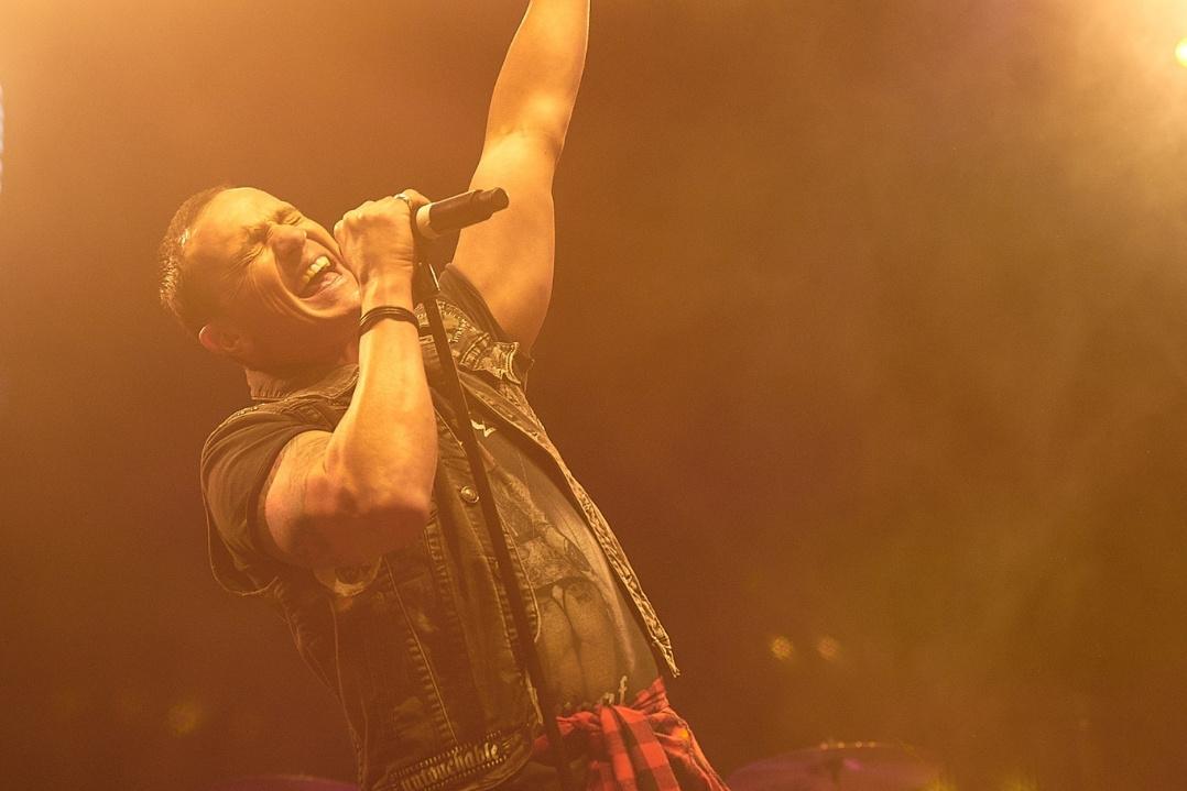 A singer passionately performs on stage, holding a microphone and reaching upwards. Warm lights and smoke create an energetic, vibrant atmosphere.