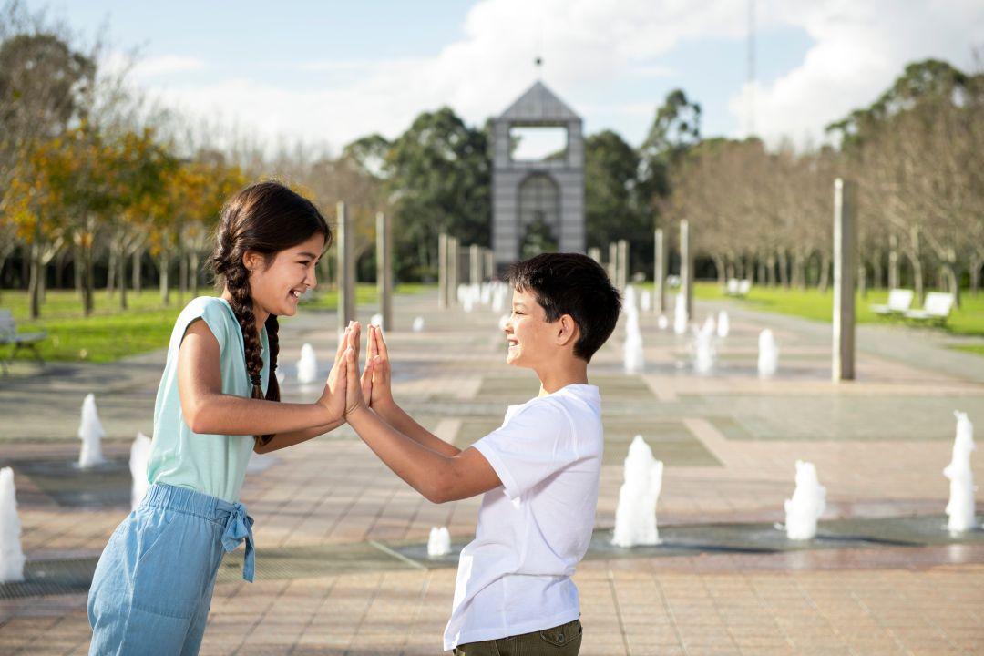 A young boy kneels, proposing playfully to a girl in a park with a monument and trees in the background. Both are smiling, creating a joyful scene.