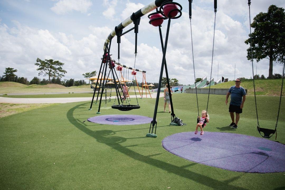 A modern playground with curved swings on a green lawn is shown under a cloudy blue sky. People play and relax, creating a joyful and lively atmosphere.