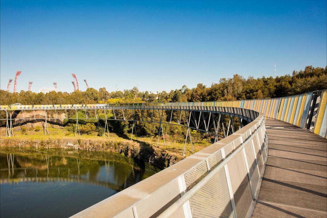 A serene wooden boardwalk curves over a calm lake, surrounded by lush green trees under a clear blue sky, conveying a peaceful, natural setting.