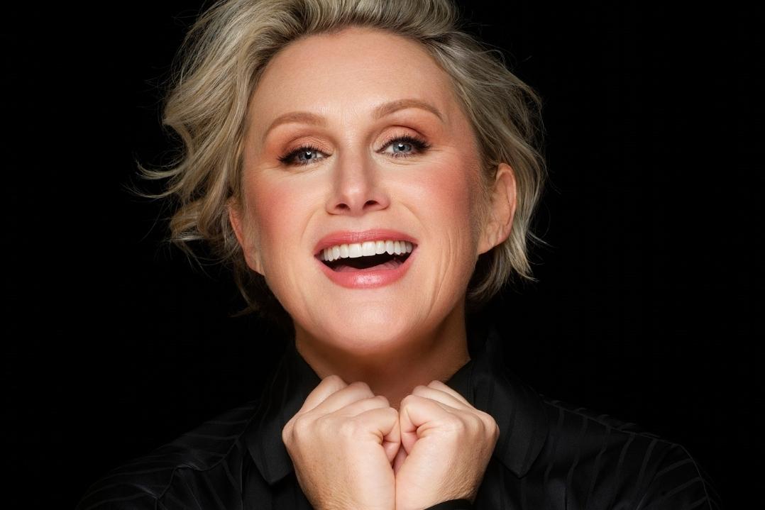 A person with short, blonde hair smiles broadly against a dark background. Their hands are clasped near their chin, conveying joy and energy.