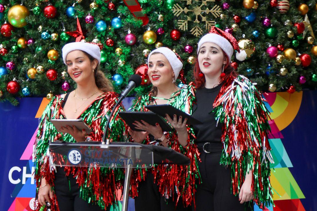 Three women, wearing festive attire and Santa hats, sing at a podium in front of a vibrant Christmas tree adorned with colourful ornaments. The scene conveys holiday cheer.