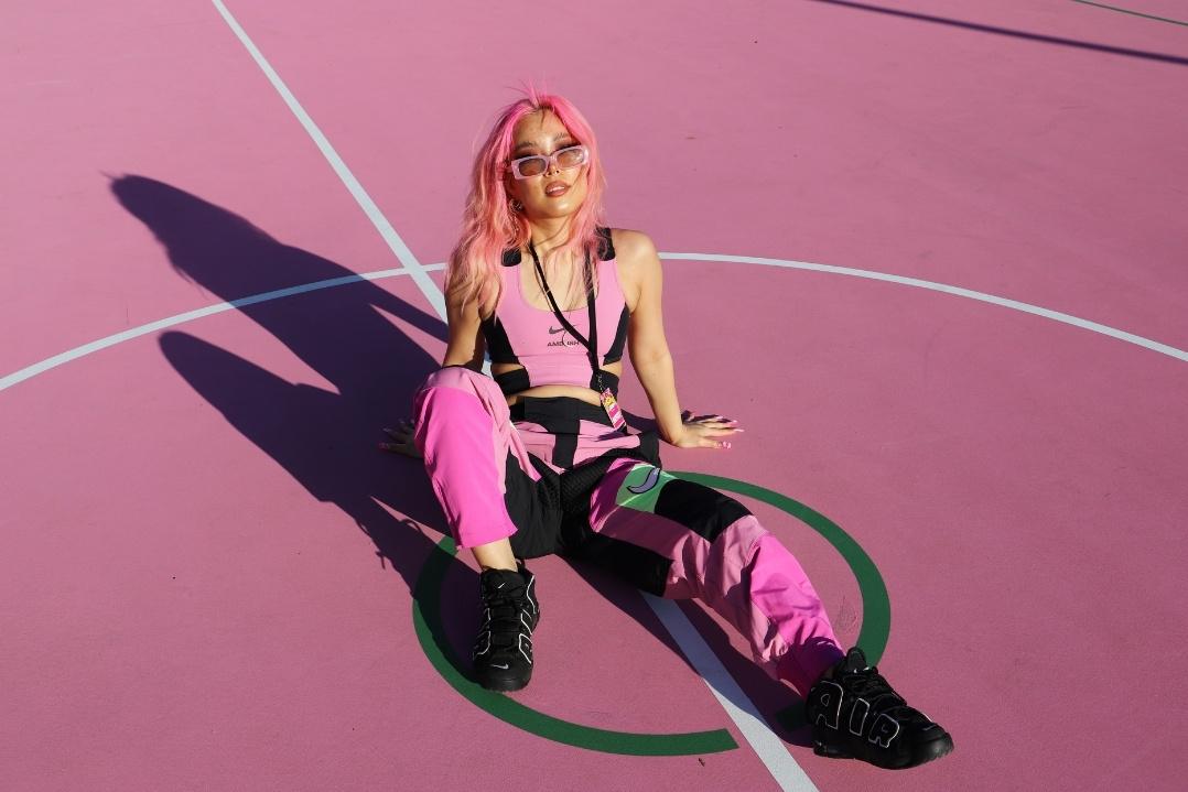 A person with pink hair and outfit sits on a pink basketball court, surrounded by roller skates. The mood is playful and stylish.